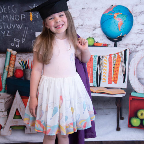 smiling girl with cap and gown wearing many places you go dress in front of a school backdrop