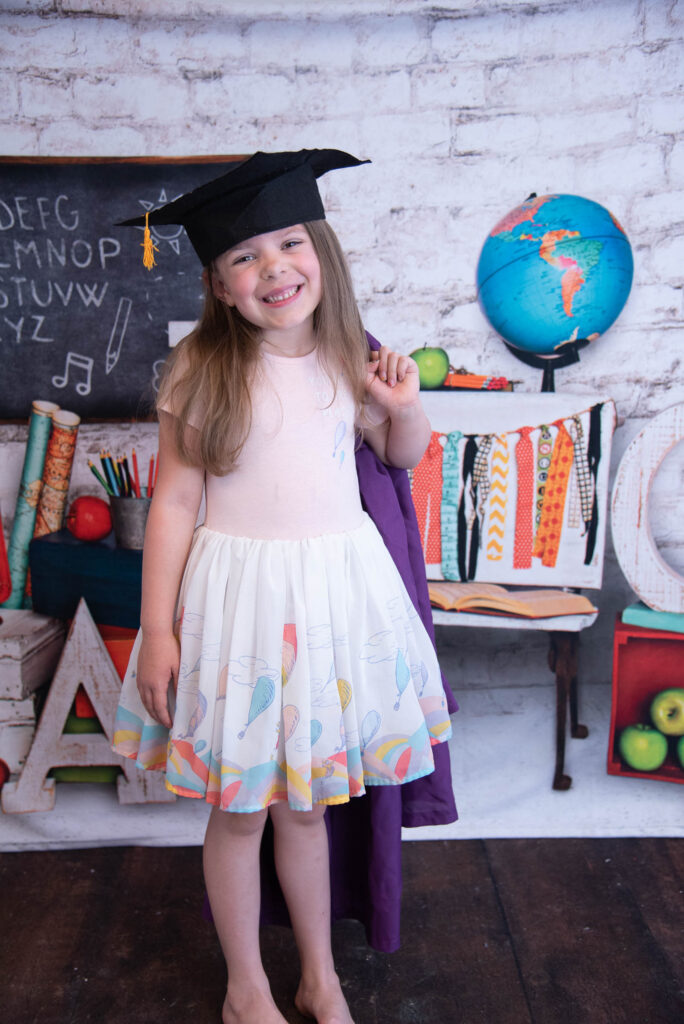 smiling girl with cap and gown wearing many places you go dress in front of a school backdrop